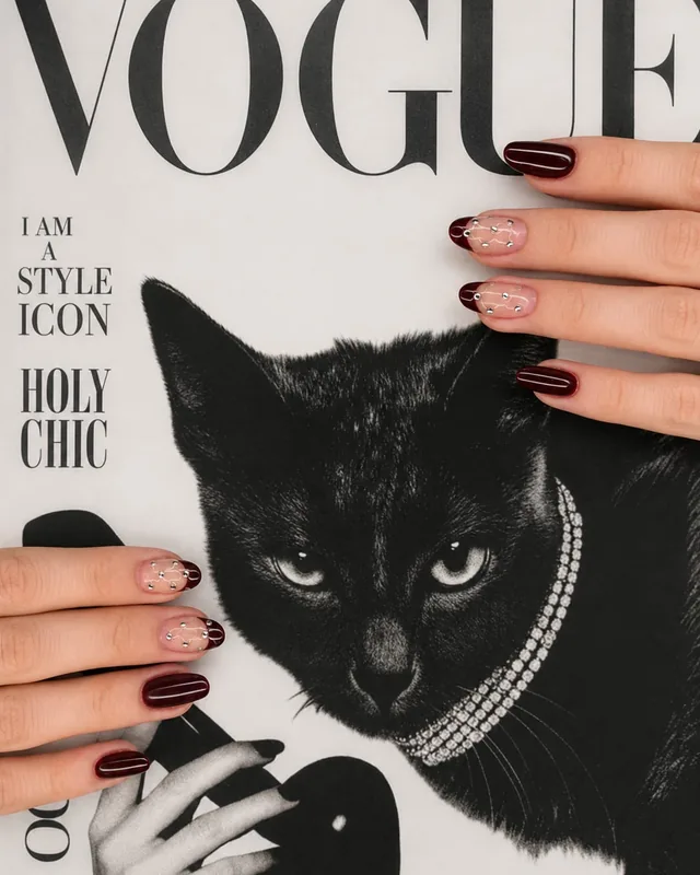 Unhas amendoadas em bordô profundo, duas delas com efeito acolchoado e cristais, sobre capa de revista com gato preto