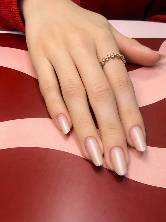 Mão com unhas amendoadas nude peroladas e anel dourado, sobre fundo ondulado em vermelho e rosa