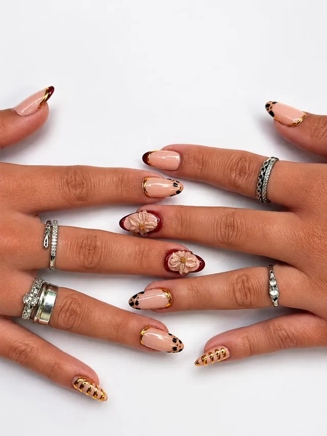 Duas mãos com unhas amendoadas nude, pontas de oncinha com filete dourado e flores em 3D, com anéis