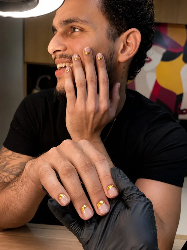 Unhas nude com pontas douradas em cliente, com a mão segurada pela profissional de luvas pretas no estúdio